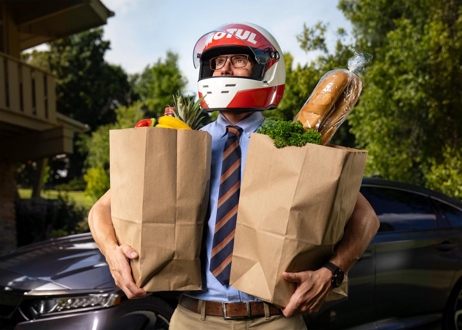 Average working man in a business outfit wears a racing helmet with the Motul logo painted ont he front. He's carrying in groceries from his Asian Import car. Proud, happy look on his face.